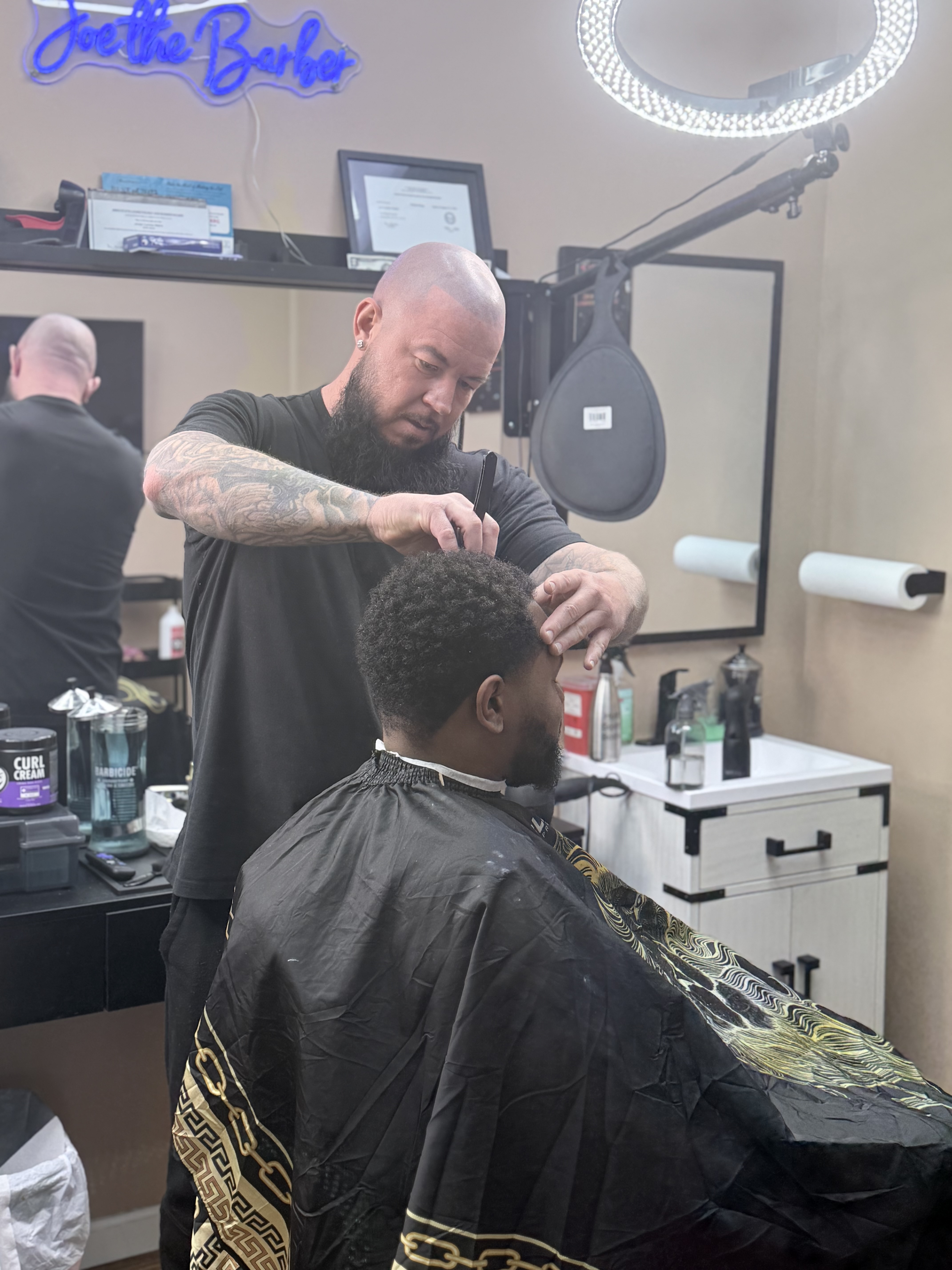 Joe Currence — owner of Joe's Barber Studio, working on a client with his signature neon sign in the background